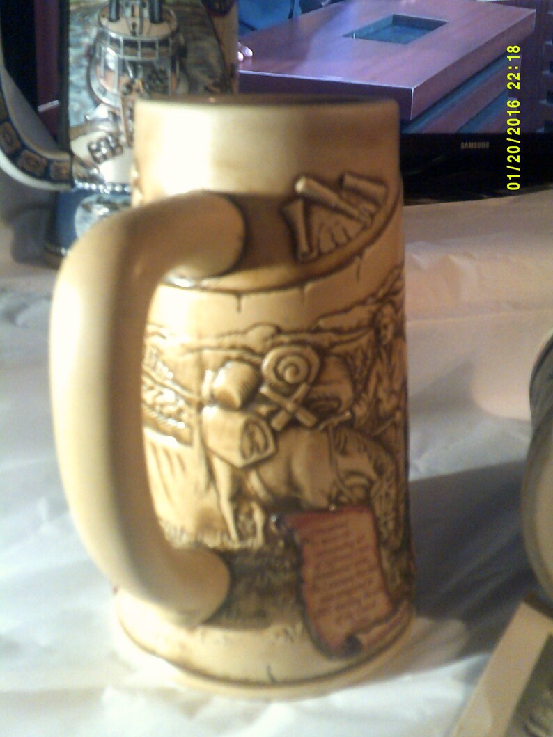 May include: A beige ceramic beer stein with a handle. The stein features a detailed relief design of a scene with people and horses. The design is in shades of brown and beige.