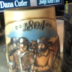 May include: A ceramic stein with a brown, tan, and white design. The stein features a scene of three men in period clothing. The year 1804 is written on the stein.