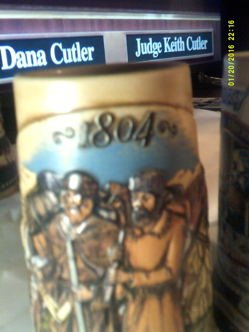 May include: A ceramic stein with a brown, tan, and white design. The stein features a scene of three men in period clothing. The year 1804 is written on the stein.