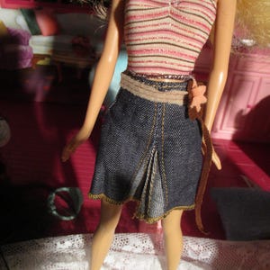 May include: A doll wearing a striped pink and white tube top with a dark halter strap. The doll is also wearing a denim skirt with a pleated front and a brown belt with a flower accent. The skirt has gold stitching.