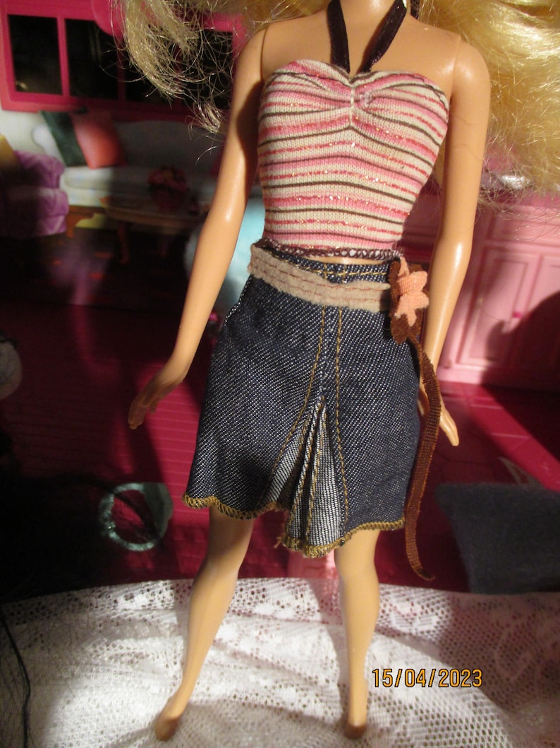 May include: A doll wearing a striped pink and white tube top with a dark halter strap. The doll is also wearing a denim skirt with a pleated front and a brown belt with a flower accent. The skirt has gold stitching.