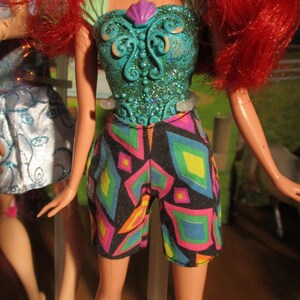 Puede incluir: Mu&ntilde;eca con cabello rojo, vestida con un top turquesa con dise&ntilde;o de concha y pantalones cortos estampados. Los pantalones cortos tienen un fondo negro con formas geom&eacute;tricas en rosa, amarillo, azul y verde. El fondo est&aacute; borroso.