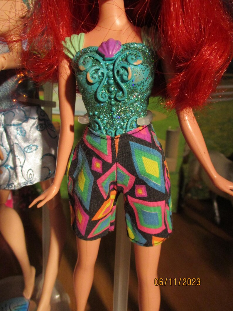 Puede incluir: Mu&ntilde;eca con cabello rojo, vestida con un top turquesa con dise&ntilde;o de concha y pantalones cortos estampados. Los pantalones cortos tienen un fondo negro con formas geom&eacute;tricas en rosa, amarillo, azul y verde. El fondo est&aacute; borroso.