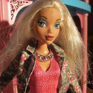 Könnte beinhalten: Eine Puppe mit langem blonden Haar, blauen Augen und grünem Lidschatten. Die Puppe trägt eine silberfarbene Halskette mit Stacheln, ein strukturiertes rosa Oberteil und eine grüne Jacke mit rosa Blumenmuster. Das Datum 15/04/20 steht unten rechts.