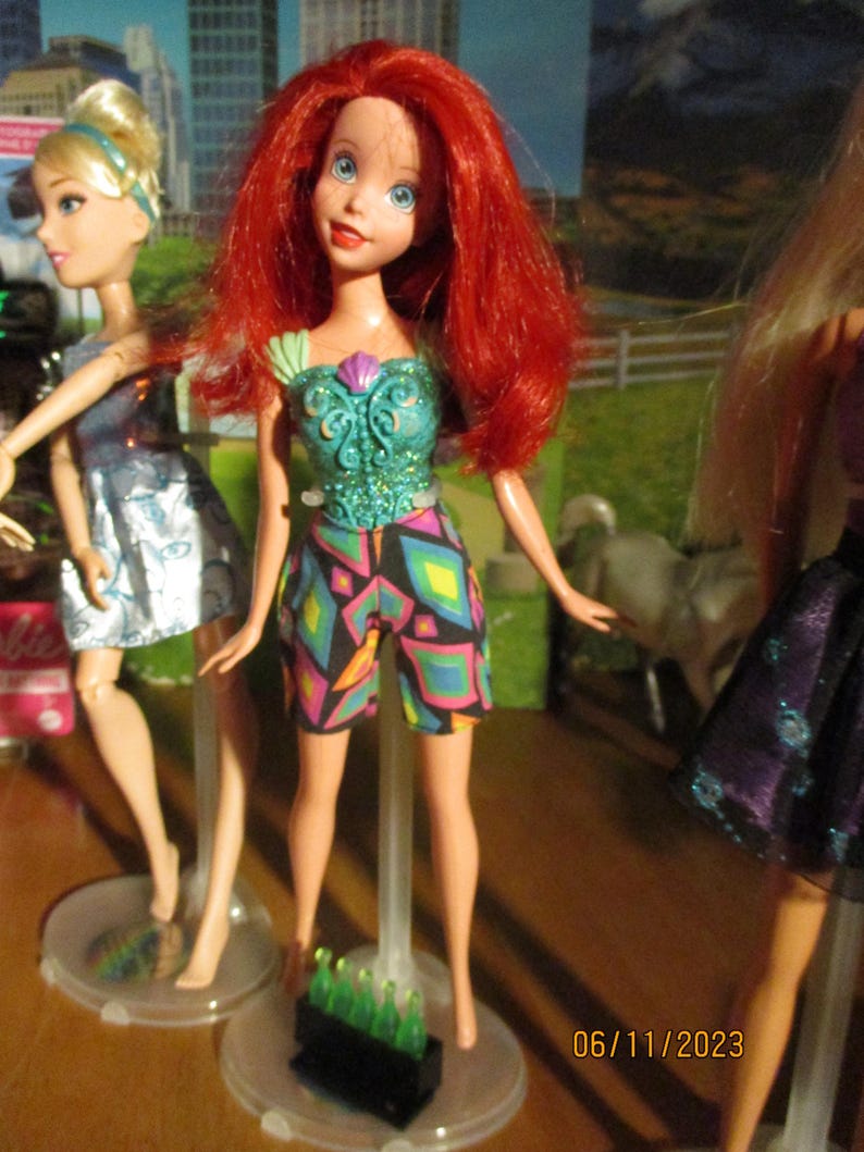 Puede incluir: Una mu&ntilde;eca con cabello rojo, ojos azules y una parte superior turquesa con un dise&ntilde;o de concha. La mu&ntilde;eca lleva pantalones cortos coloridos. Otras mu&ntilde;ecas son visibles en el fondo. La fecha 06/11/2023 est&aacute; impresa en la esquina inferior derecha.