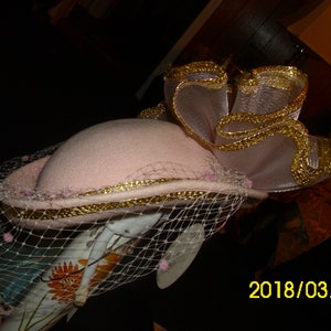May include: A pink hat with a gold trim and a white veil. The hat has a large, ruffled bow on top, also trimmed with gold.