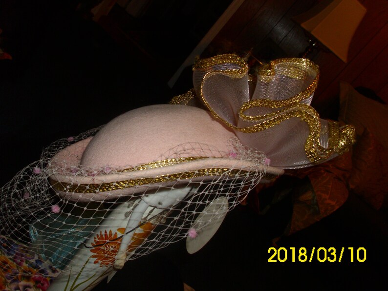 May include: A pink hat with a gold trim and a white veil. The hat has a large, ruffled bow on top, also trimmed with gold.