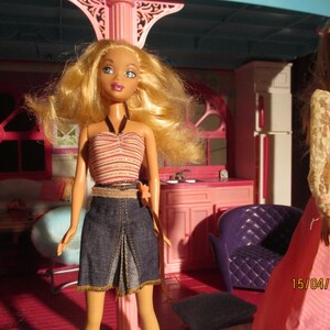May include: A doll with blonde hair wearing a striped halter top and denim skirt stands in a pink dollhouse. Another doll in a pink dress is also visible. The dollhouse has pink furniture and decor.
