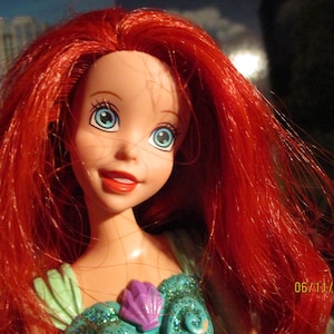 Puede incluir: Mu&ntilde;eca con cabello rojo y ojos azules, vestida con un atuendo turquesa y morado. La mu&ntilde;eca tiene una expresi&oacute;n sonriente y es un personaje de una pel&iacute;cula de animaci&oacute;n popular. La fecha 06/11/2023 es visible.