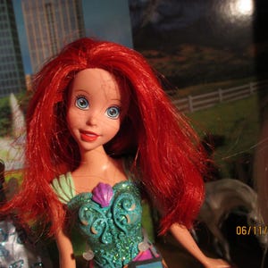 Puede incluir: Una mu&ntilde;eca con cabello rojo brillante y ojos azules, vestida con un vestido turquesa y morado. La mu&ntilde;eca tiene una expresi&oacute;n sonriente. El fondo incluye un paisaje urbano y una imagen borrosa de un caballo.
