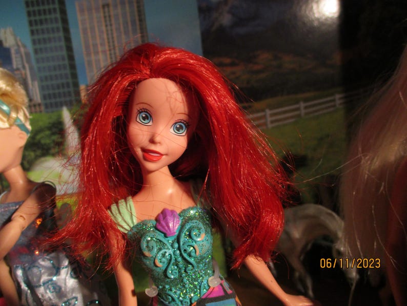 Puede incluir: Una mu&ntilde;eca con cabello rojo brillante y ojos azules, vestida con un vestido turquesa y morado. La mu&ntilde;eca tiene una expresi&oacute;n sonriente. El fondo incluye un paisaje urbano y una imagen borrosa de un caballo.
