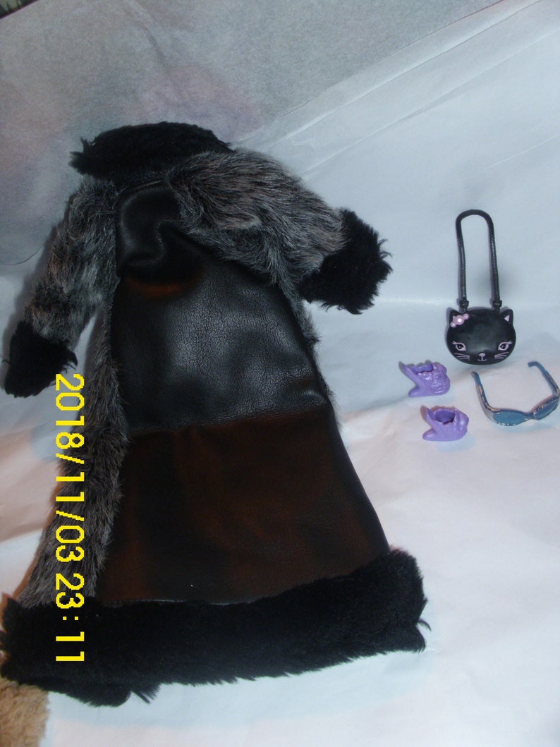 May include: A black faux fur coat with a black leather bodice and a black fur trim. The coat is laying on a white surface with a black cat purse, purple gloves, and sunglasses.