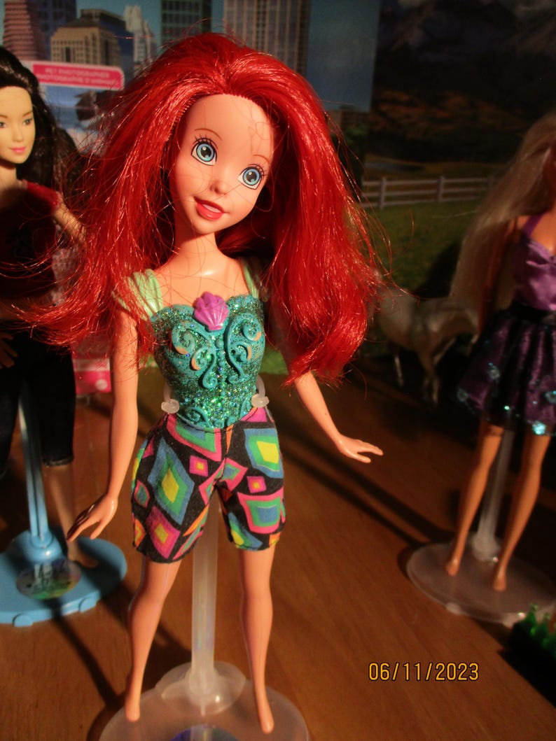 Puede incluir: Mu&ntilde;eca con cabello rojo, ojos azules y un top turquesa con una concha rosa. La mu&ntilde;eca lleva pantalones cortos con estampado geom&eacute;trico colorido. Otras mu&ntilde;ecas son visibles en el fondo.