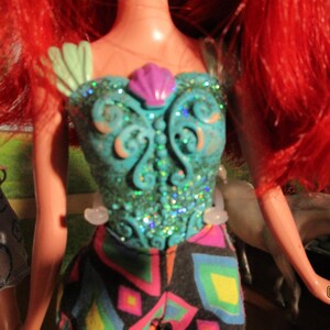 Puede incluir: Una mu&ntilde;eca con cabello rojo brillante y un corpi&ntilde;o turquesa con un dise&ntilde;o de concha. El corpi&ntilde;o est&aacute; adornado con purpurina y un acento de concha rosa. La mu&ntilde;eca lleva pantalones estampados con una variedad de colores.