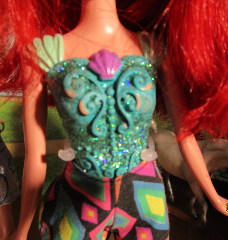 Puede incluir: Una mu&ntilde;eca con cabello rojo brillante y un corpi&ntilde;o turquesa con un dise&ntilde;o de concha. El corpi&ntilde;o est&aacute; adornado con purpurina y un acento de concha rosa. La mu&ntilde;eca lleva pantalones estampados con una variedad de colores.