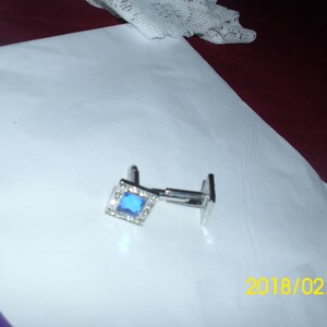 May include: A pair of silver cufflinks with a blue square gemstone surrounded by smaller clear gemstones.