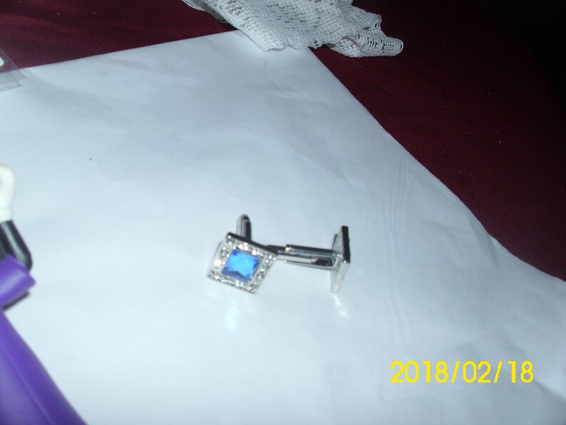 May include: A pair of silver cufflinks with a blue square gemstone surrounded by smaller clear gemstones.