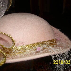 May include: A pink hat with a gold trim and a white veil. The veil has small pink flowers attached to it. The hat is decorated with a gold and white ribbon.