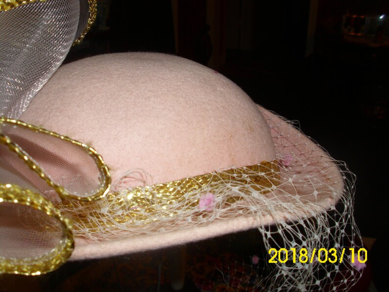 May include: A pink hat with a gold trim and a white veil. The veil has small pink flowers attached to it. The hat is decorated with a gold and white ribbon.