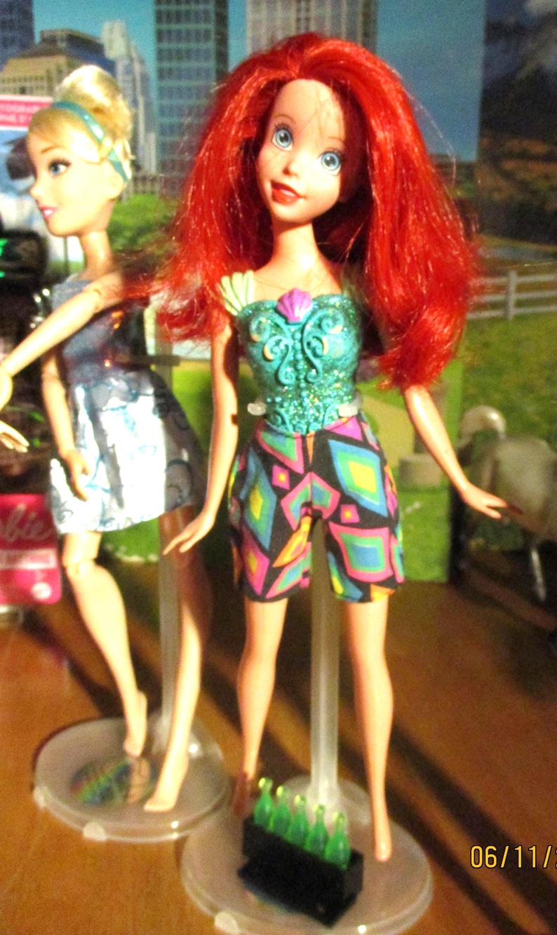 Puede incluir: Se muestran dos mu&ntilde;ecas de moda. Una mu&ntilde;eca tiene el pelo rojo y lleva un top turquesa y pantalones cortos estampados. La otra mu&ntilde;eca tiene el pelo rubio y un vestido azul. Ambas mu&ntilde;ecas est&aacute;n sobre soportes transparentes.