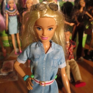 May include: A blonde doll wearing a blue denim shirt dress with a red and white striped belt. The doll has blue eyes and is wearing sunglasses.