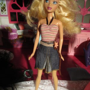 May include: A fashion doll with blonde hair and blue eyes, wearing a striped top, denim skirt, and brown belt. The doll is posed in front of a pink dollhouse interior. The date 15/04/2023 is visible in the bottom right corner.
