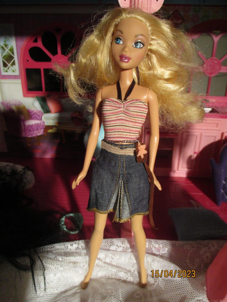 May include: A fashion doll with blonde hair and blue eyes, wearing a striped top, denim skirt, and brown belt. The doll is posed in front of a pink dollhouse interior. The date 15/04/2023 is visible in the bottom right corner.