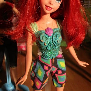 Puede incluir: Mu&ntilde;eca con cabello rojo y ojos azules, vestida con un top turquesa con dise&ntilde;o de concha y pantalones cortos coloridos. La mu&ntilde;eca est&aacute; sobre un soporte transparente, con otra mu&ntilde;eca visible al fondo.