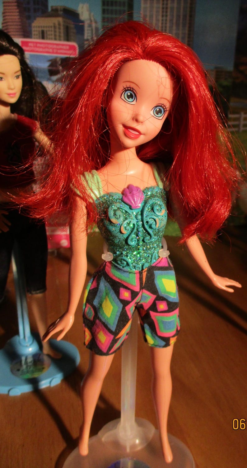 Puede incluir: Mu&ntilde;eca con cabello rojo y ojos azules, vestida con un top turquesa con dise&ntilde;o de concha y pantalones cortos coloridos. La mu&ntilde;eca est&aacute; sobre un soporte transparente, con otra mu&ntilde;eca visible al fondo.