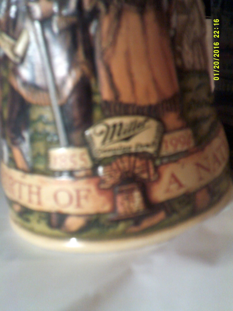 May include: A brown and white ceramic stein with a green, brown, and white design. The stein features a banner with the text "1965" and "1995" and the words "Birth of A Nation". The stein also features a logo for Miller Brewing Company.