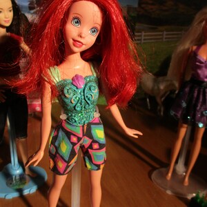 Puede incluir: Mu&ntilde;eca con cabello rojo, ojos azules y un top turquesa con una concha rosa. La mu&ntilde;eca lleva pantalones cortos con estampado geom&eacute;trico colorido. Otras mu&ntilde;ecas son visibles en el fondo.