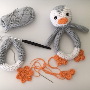 May include: A gray and white crocheted penguin rattle with orange crocheted feet. The rattle is partially assembled with a crochet hook and yarn nearby.