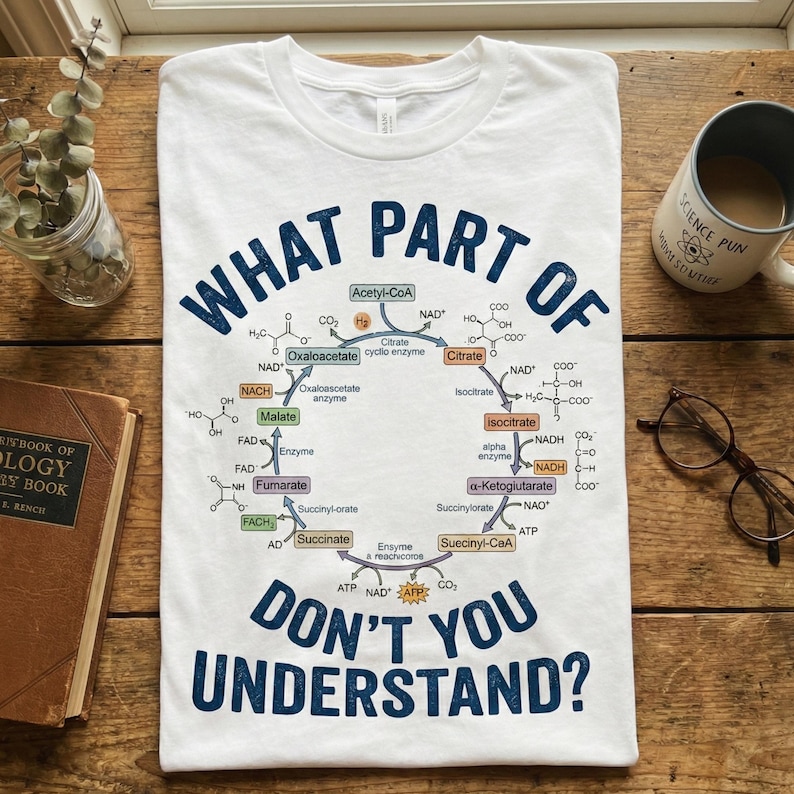 May include: White t-shirt with a blue graphic of a biochemical cycle and the text "WHAT PART OF DON'T YOU UNDERSTAND?" The shirt is displayed on a wooden surface with a coffee mug, glasses, and a book.