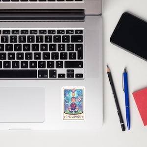 May include: A sticker featuring a tarot card design with a cartoon illustration of a person meditating. The card has a blue and purple border with the text "THE WINNER" at the bottom. The person is wearing a red and black shirt.