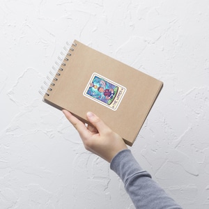 May include: A sticker featuring a colorful illustration of a person surfing on a wave, with the text "THE WINNER" around the border. The sticker is on a brown spiral notebook.