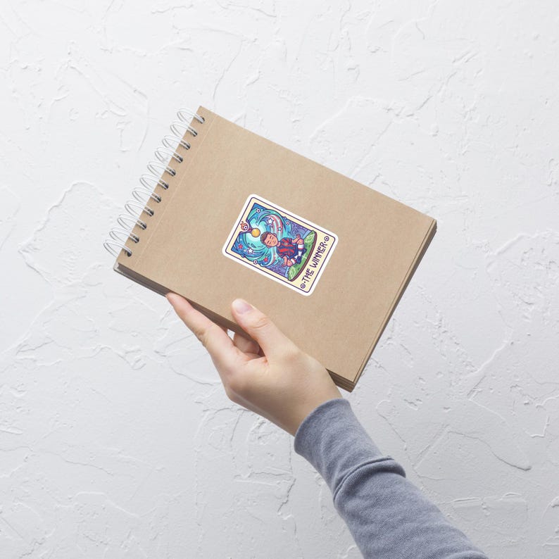 May include: A sticker featuring a colorful illustration of a person surfing on a wave, with the text "THE WINNER" around the border. The sticker is on a brown spiral notebook.