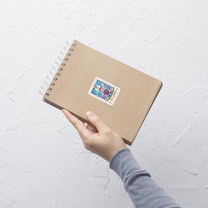 May include: A sticker featuring a tarot card design, with the words "The Lovers" at the bottom. The card depicts a figure with a halo, surrounded by colorful, swirling imagery. The sticker is on a brown spiral notebook.