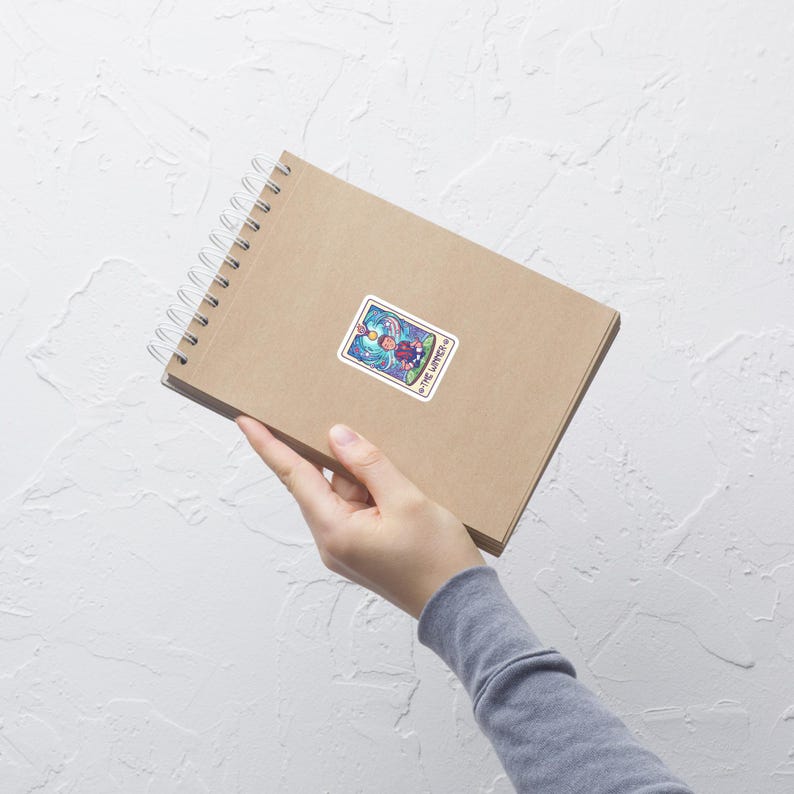 May include: A sticker featuring a tarot card design, with the words "The Lovers" at the bottom. The card depicts a figure with a halo, surrounded by colorful, swirling imagery. The sticker is on a brown spiral notebook.