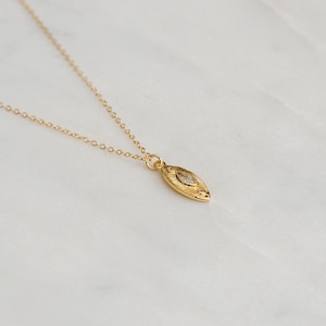 May include: A gold necklace featuring a fine chain and a pendant. The pendant is an elongated oval with a textured design and a small, clear stone. The necklace is presented on a white marble surface.