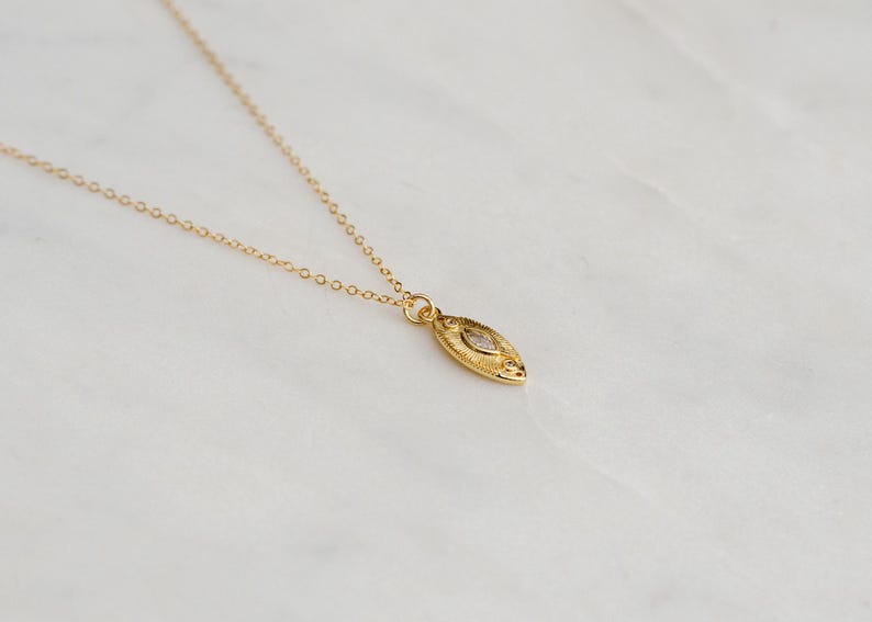 May include: A gold necklace featuring a fine chain and a pendant. The pendant is an elongated oval with a textured design and a small, clear stone. The necklace is presented on a white marble surface.