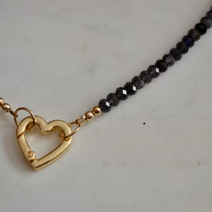 Op de afbeelding: Een ketting met een gouden hartvormige sluiting en een kralenketting. De donkergrijze, gefacetteerde kralen cre&euml;ren een sprankelend effect. De gouden sluiting voegt een vleugje elegantie toe aan de sieraden.