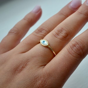 May include: A gold-colored ring featuring an eye-shaped design. The eye has a white background with a blue iris and a gold border. The ring is shown on a person's finger.