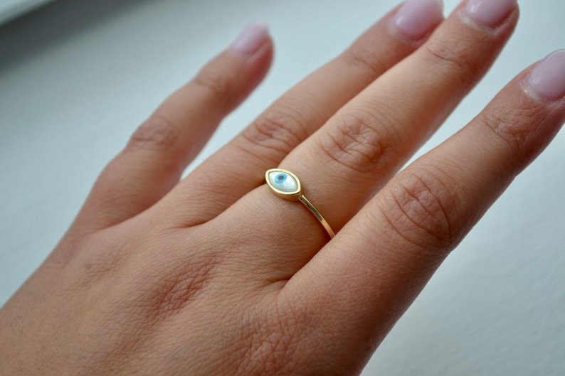 May include: A gold-colored ring featuring an eye-shaped design. The eye has a white background with a blue iris and a gold border. The ring is shown on a person's finger.