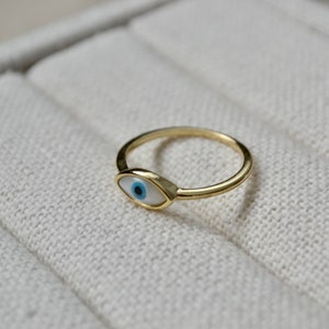 May include: A gold-colored ring featuring an eye-shaped design with a white background, a blue iris, and a black pupil. The ring is set on a light beige surface.