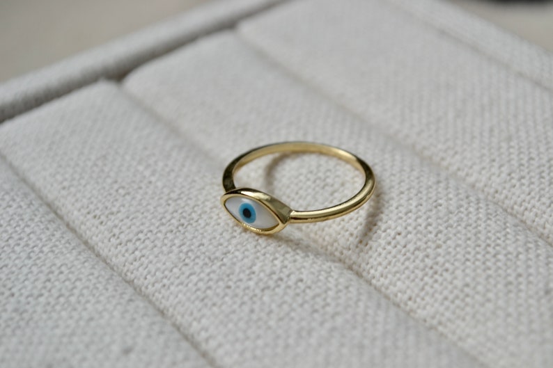 May include: A gold-colored ring featuring an eye-shaped design with a white background, a blue iris, and a black pupil. The ring is set on a light beige surface.