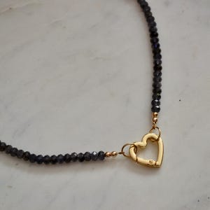 Op de afbeelding: Een ketting met donkerblauwe facetkralen en een goudkleurige hartvormige sluiting. De ketting heeft kleine goudkleurige accenten en wordt gepresenteerd op een wit marmeren oppervlak. Een delicaat sieraad.