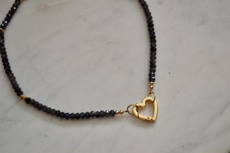 Op de afbeelding: Een ketting met donkerblauwe facetkralen en een goudkleurige hartvormige sluiting. De ketting heeft kleine goudkleurige accenten en wordt gepresenteerd op een wit marmeren oppervlak. Een delicaat sieraad.