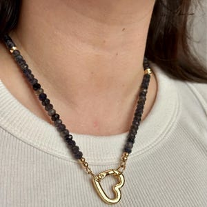 Op de afbeelding: Een ketting met donkere, gefacetteerde kralen en gouden accenten. De ketting heeft een goudkleurige hartvormige sluiting. De ketting wordt gedragen over een witte top.