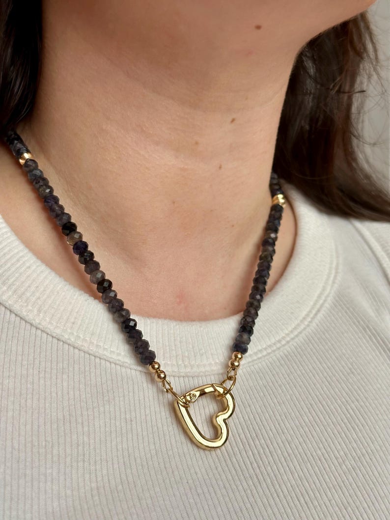 Op de afbeelding: Een ketting met donkere, gefacetteerde kralen en gouden accenten. De ketting heeft een goudkleurige hartvormige sluiting. De ketting wordt gedragen over een witte top.