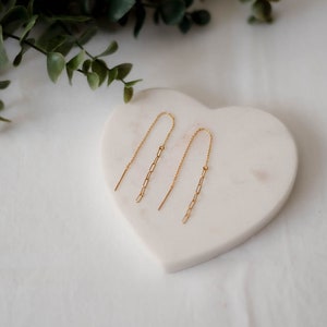 Może przedstawiać: Para złotych kolczyków z delikatnym łańcuszkiem. Kolczyki są prezentowane na białym sercu.